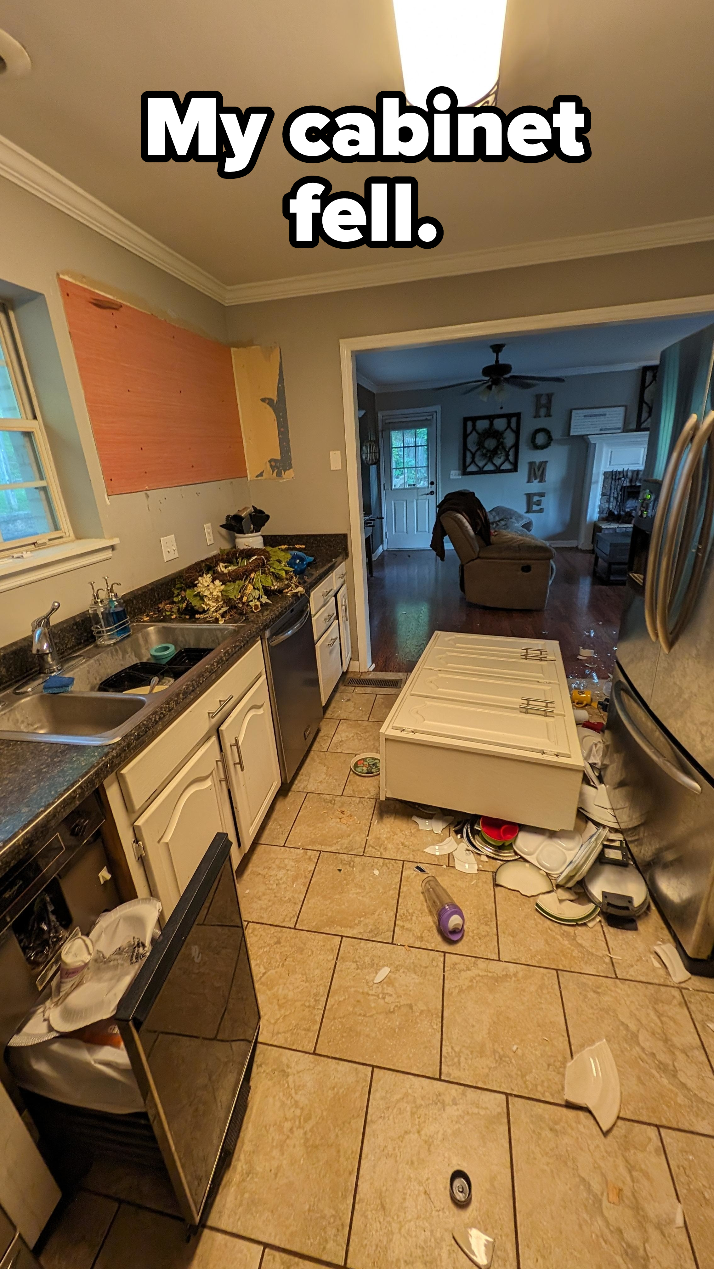 A kitchen with a broken fridge door lying on the floor, scattered items, and debris. The living room in the background appears unaffected. No people are present