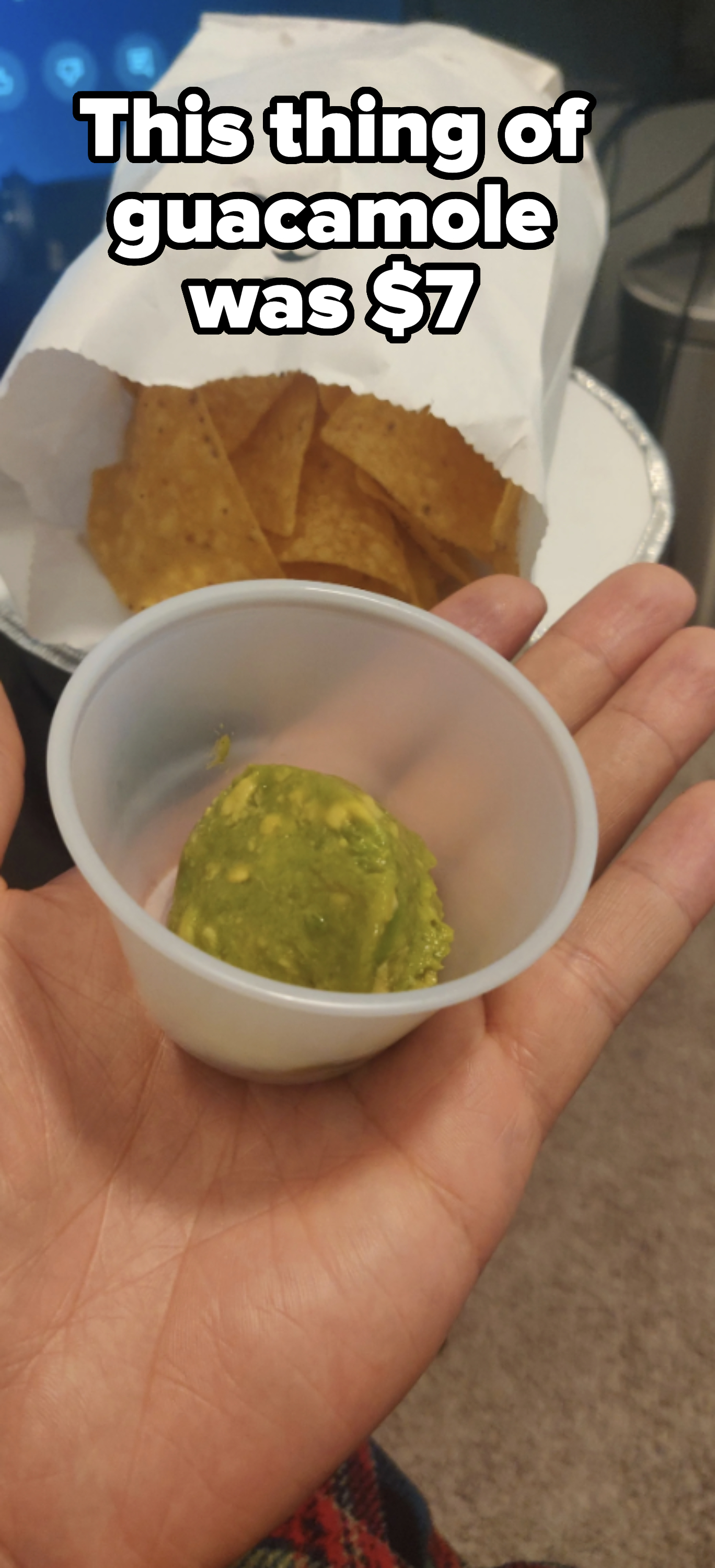 A hand holds a small cup of guacamole in front of a plate with a paper bag containing tortilla chips