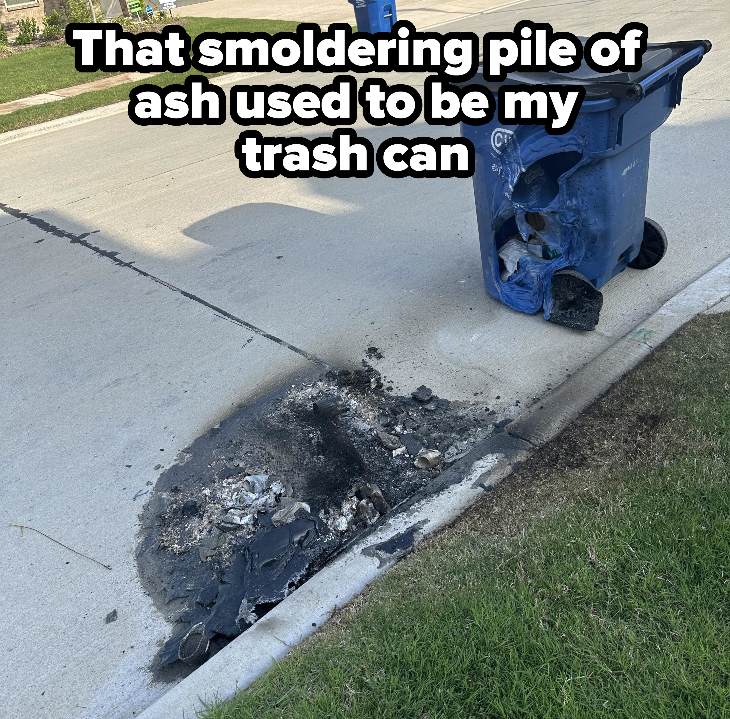 A melted and burned blue trash bin on a suburban street, with a charred area on the pavement next to it