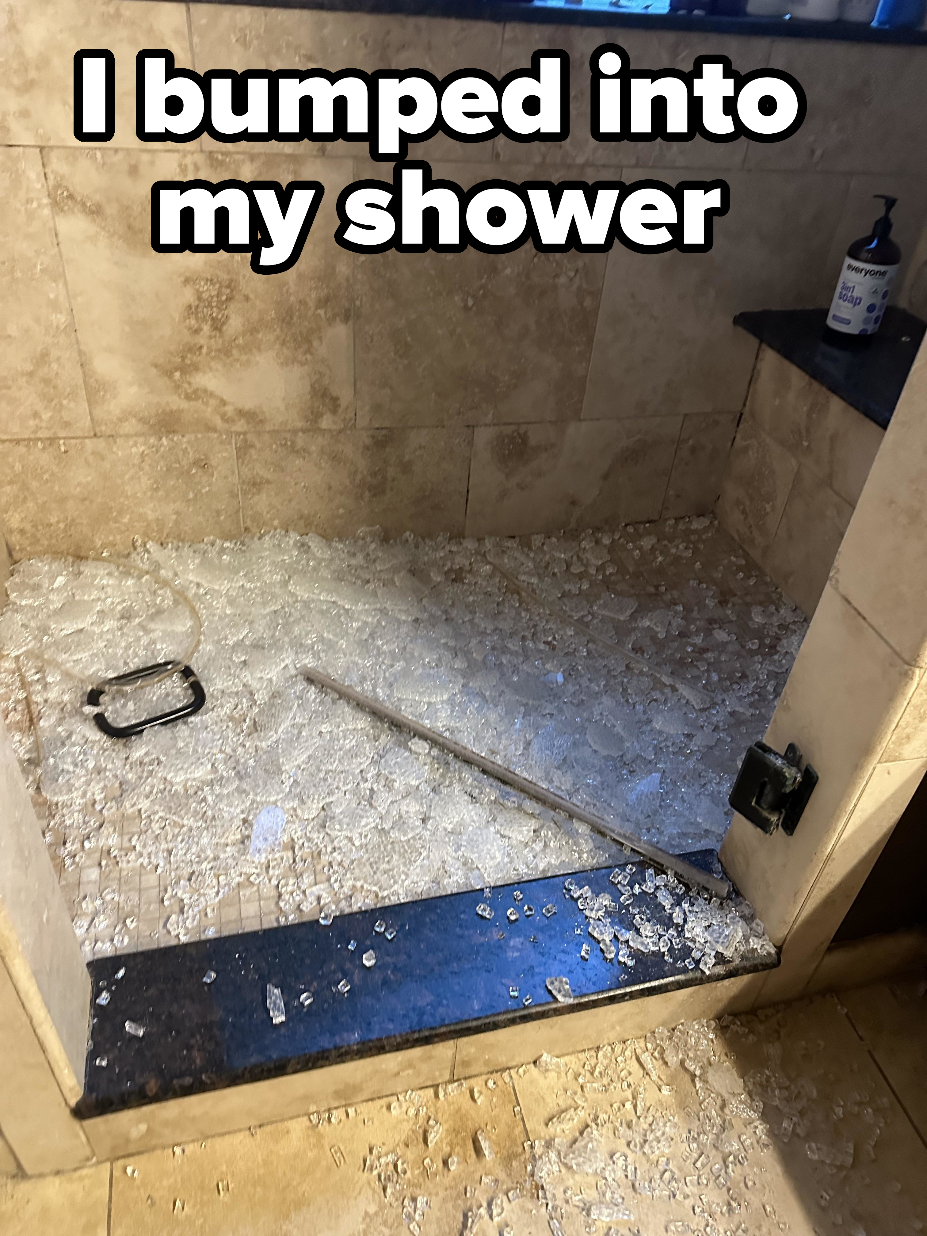 A shattered glass shower door lies in pieces on the shower floor, with small fragments scattered all around. A bottle of soap is visible on the ledge