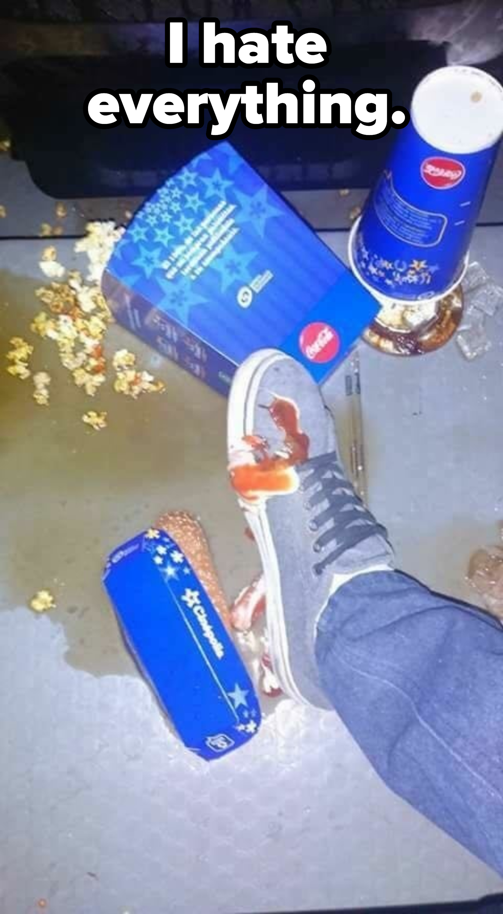 A spilled drink and popcorn at a movie theater, with a foot in a sneaker stepping on some of the mess