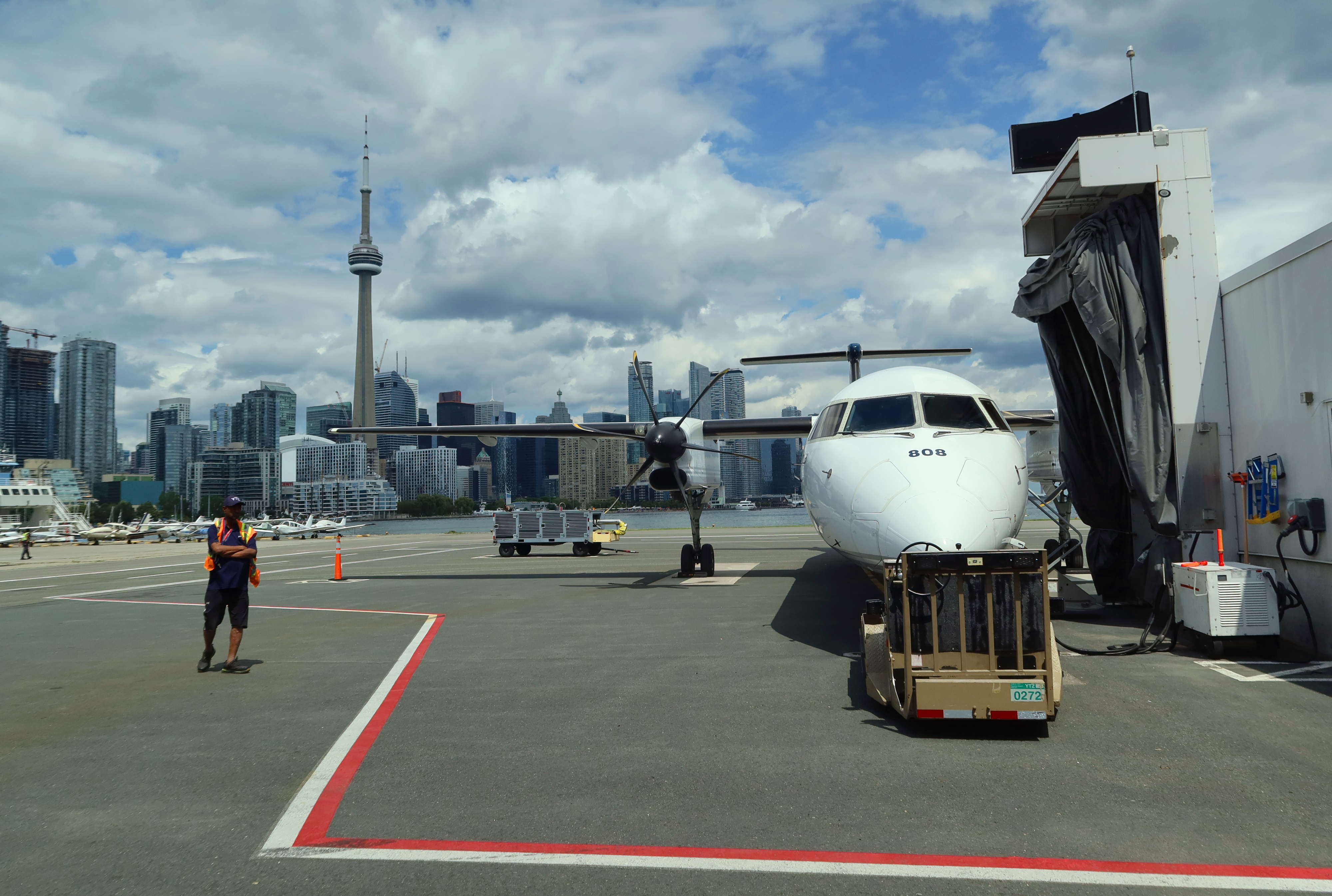 An airplane is parked at an airport gate. A worker is walking on the tarmac. The city skyline, including a tall tower, is visible in the background