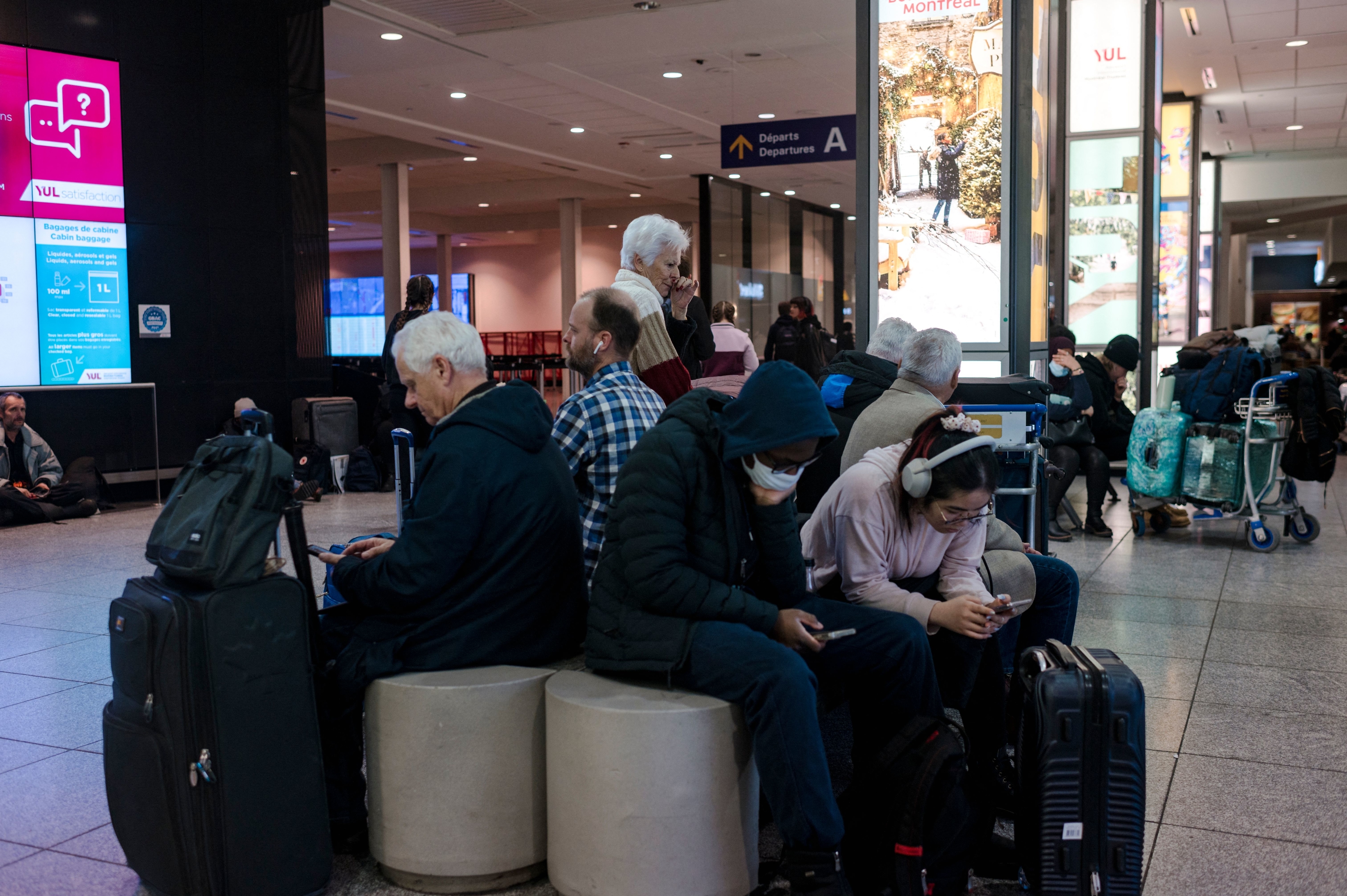 Travelers in an airport terminal sitting on circular benches, with luggage around them, using their phones and waiting. Signs and screens are visible in the background