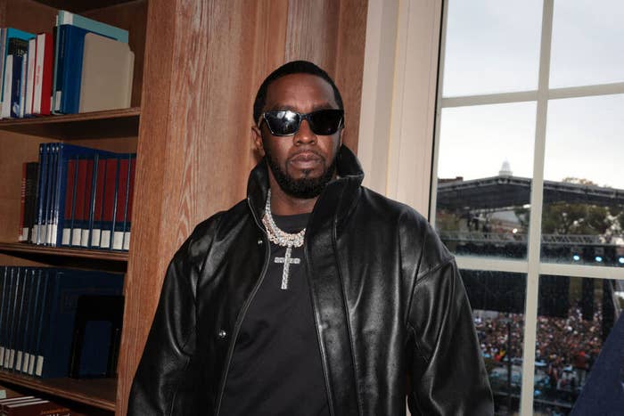 Sean &quot;Diddy&quot; Combs wearing a black leather jacket and sunglasses, standing indoors with a bookshelf behind him and a crowd outside a window