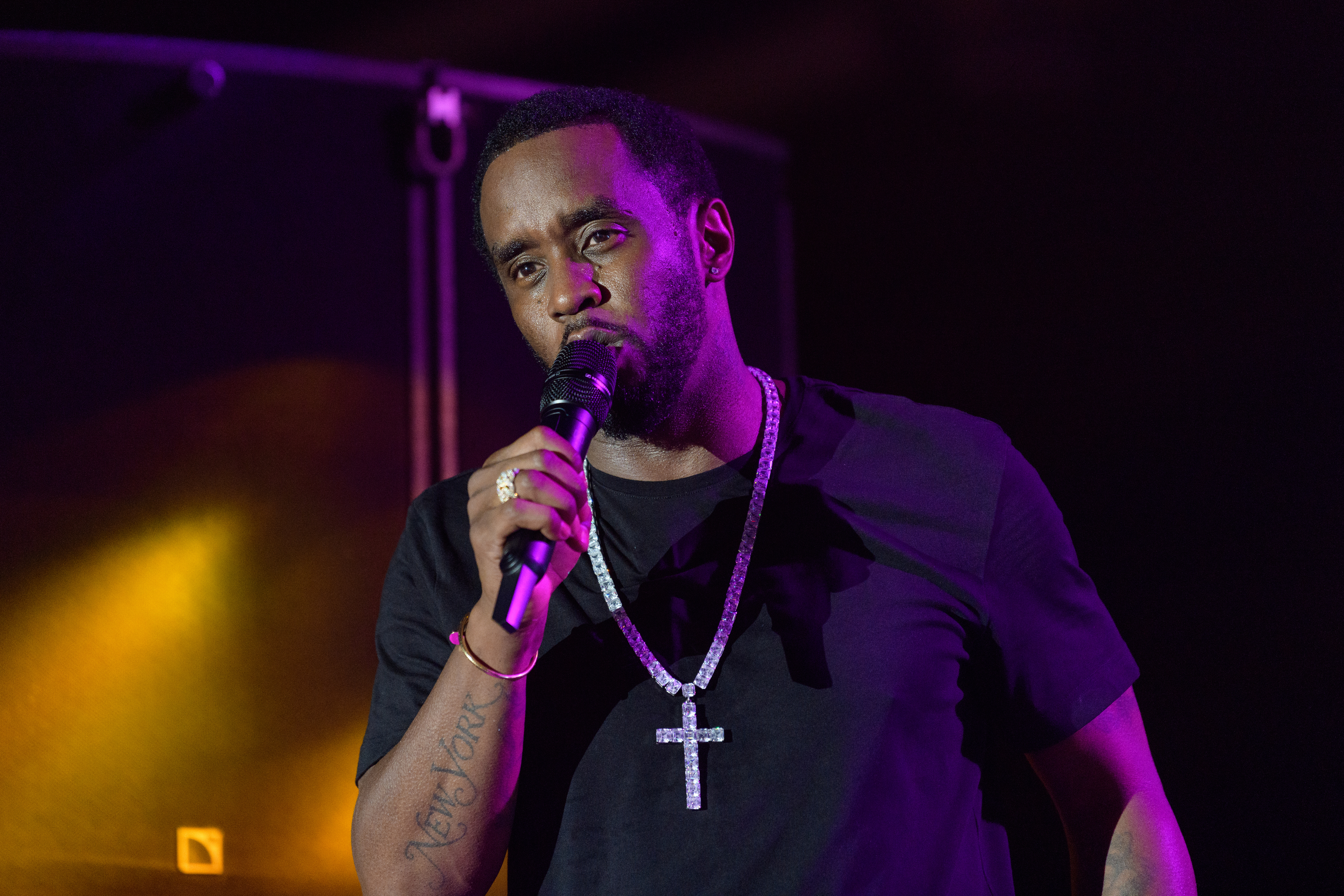 Sean "Diddy" Combs holds a microphone on stage, wearing a black shirt and a large jeweled cross necklace