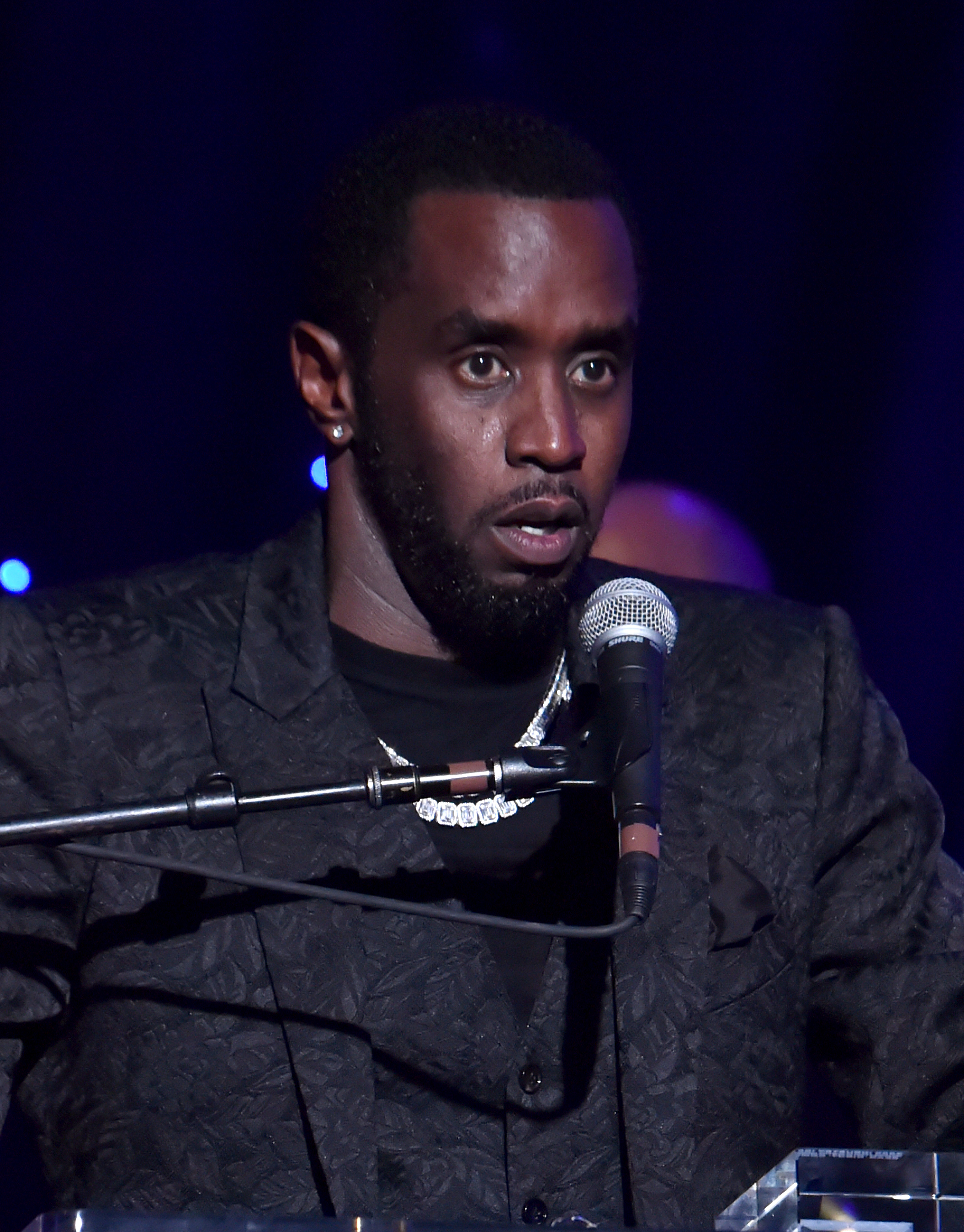 Sean &quot;Diddy&quot; Combs, dressed in a textured suit and chain necklace, speaking into a microphone at an event