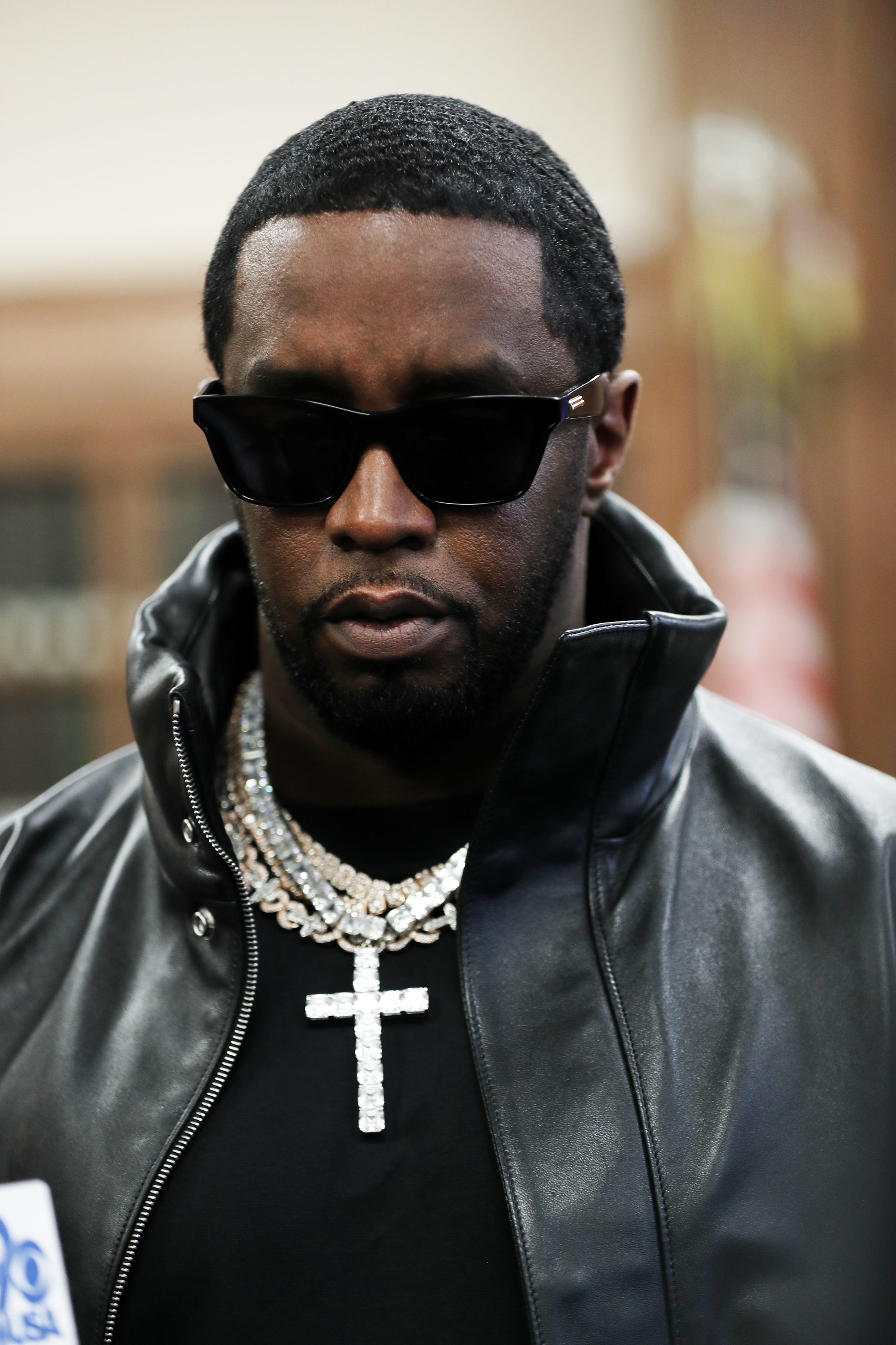 Sean Combs wears a leather jacket, sunglasses, and layered necklaces with one cross pendant. He stands in a formal setting