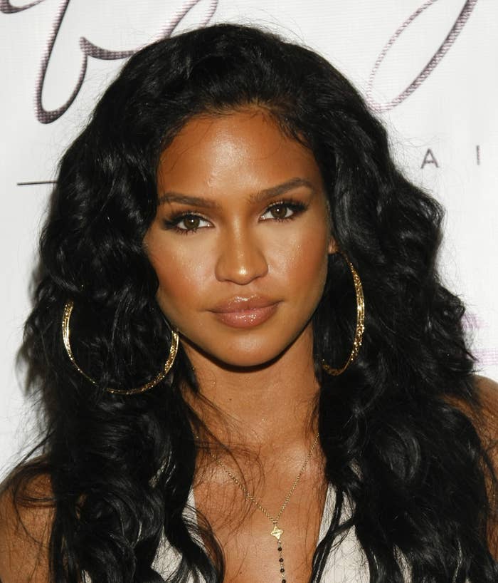 Cassie Ventura poses in front of a backdrop with wavy hair, large hoop earrings, and a necklace