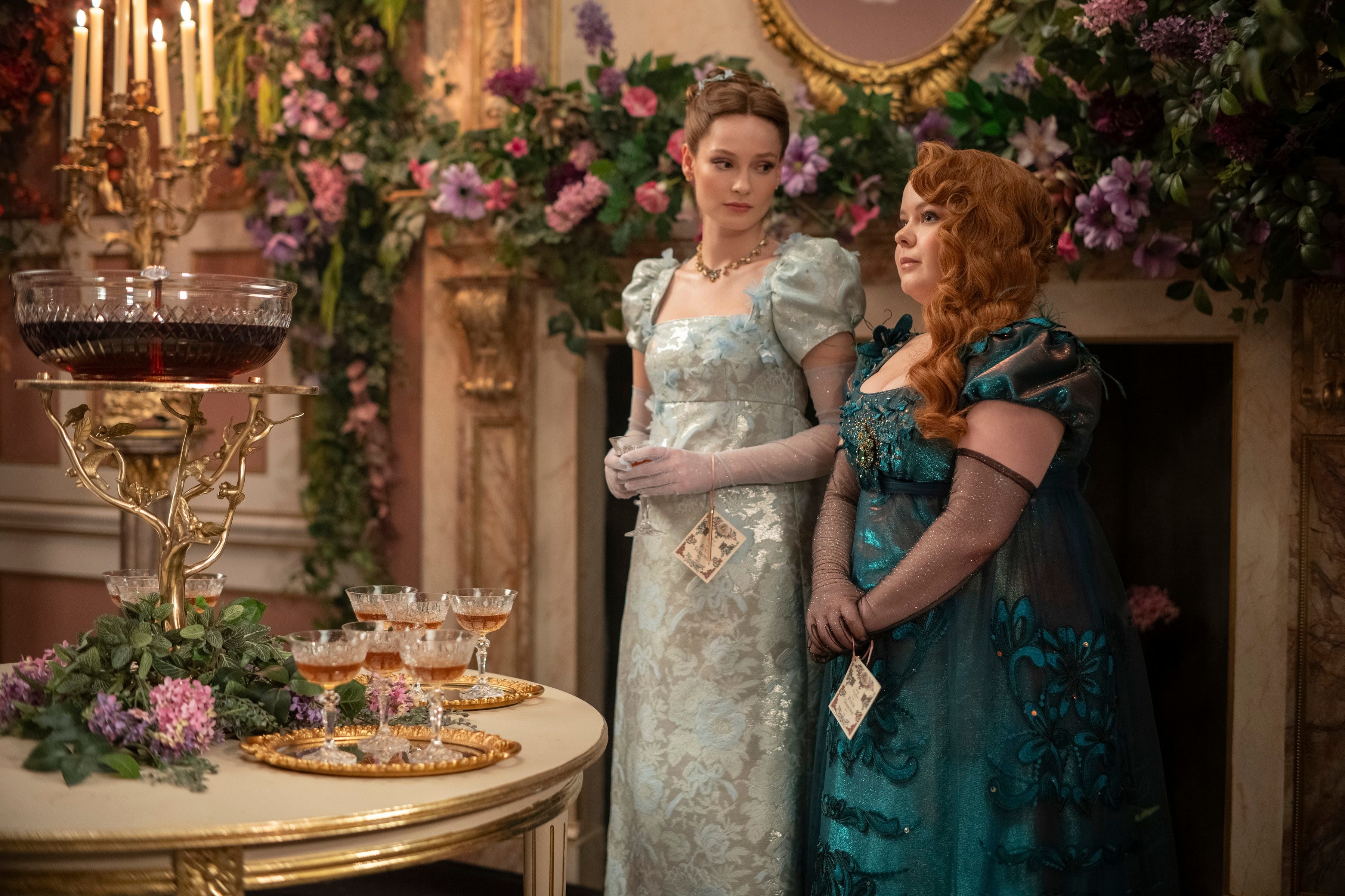 Phoebe Dynevor and Nicola Coughlan in elegant, regency-style gowns, standing next to a table with drinks, surrounded by lush floral decorations