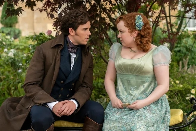 Jonathon Bailey and Nicola Coughlan in period costume. Jonathon wears a brown coat and Nicola a green dress with sheer sleeves and a floral hair accessory