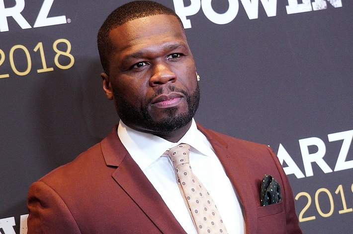 50 Cent at an event for Starz's "Power" in 2018, wearing a brown suit with a patterned tie and pocket square. The background features the event's branding