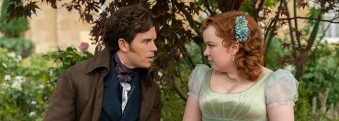 Luke Newton and Nicola Coughlan in period costume. Luke wears a brown coat and Nicola a green dress with sheer sleeves and a floral hair accessory