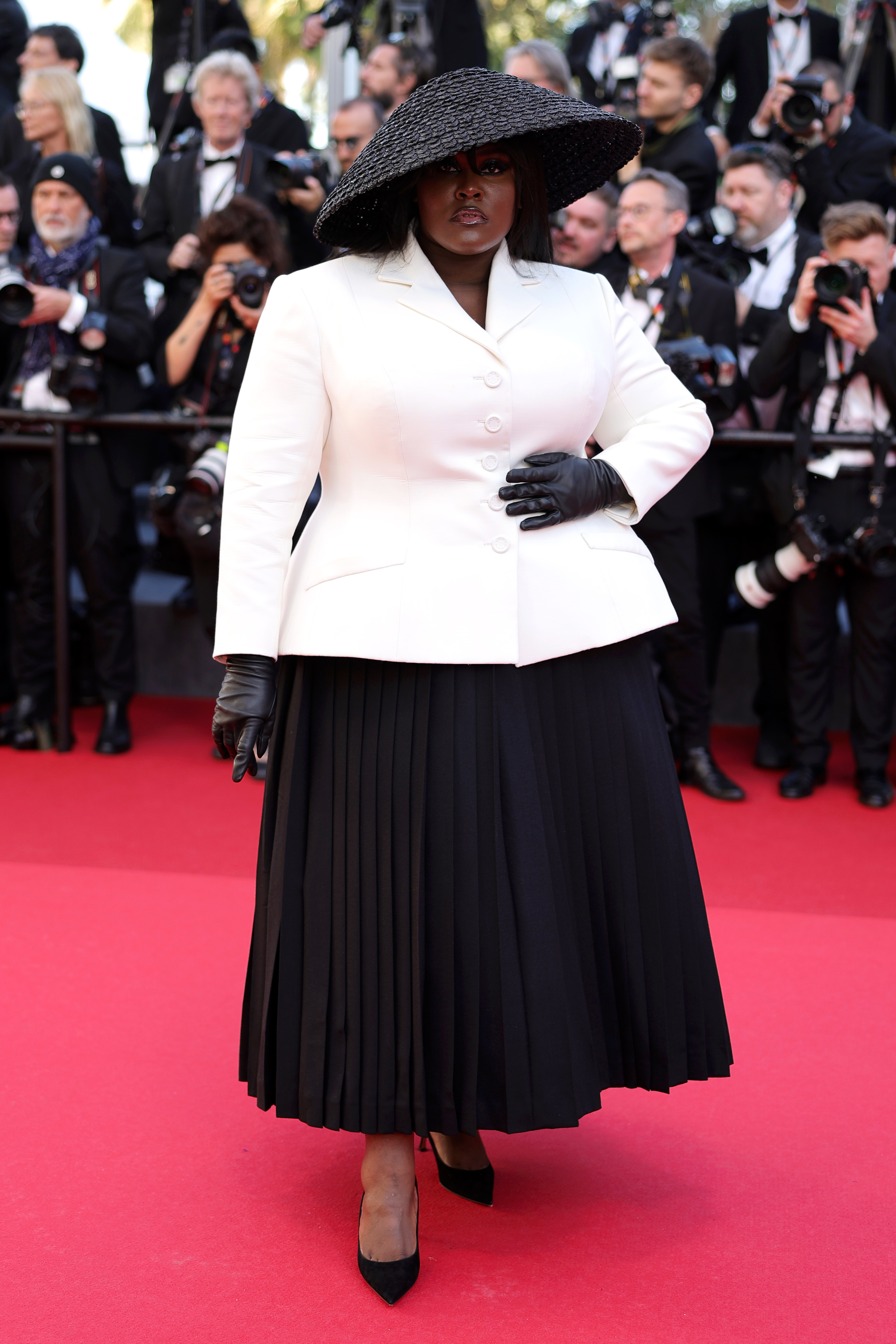 Yseult on the red carpet, wearing a white blazer, black pleated skirt, oversized black hat, and black gloves, posing for photos