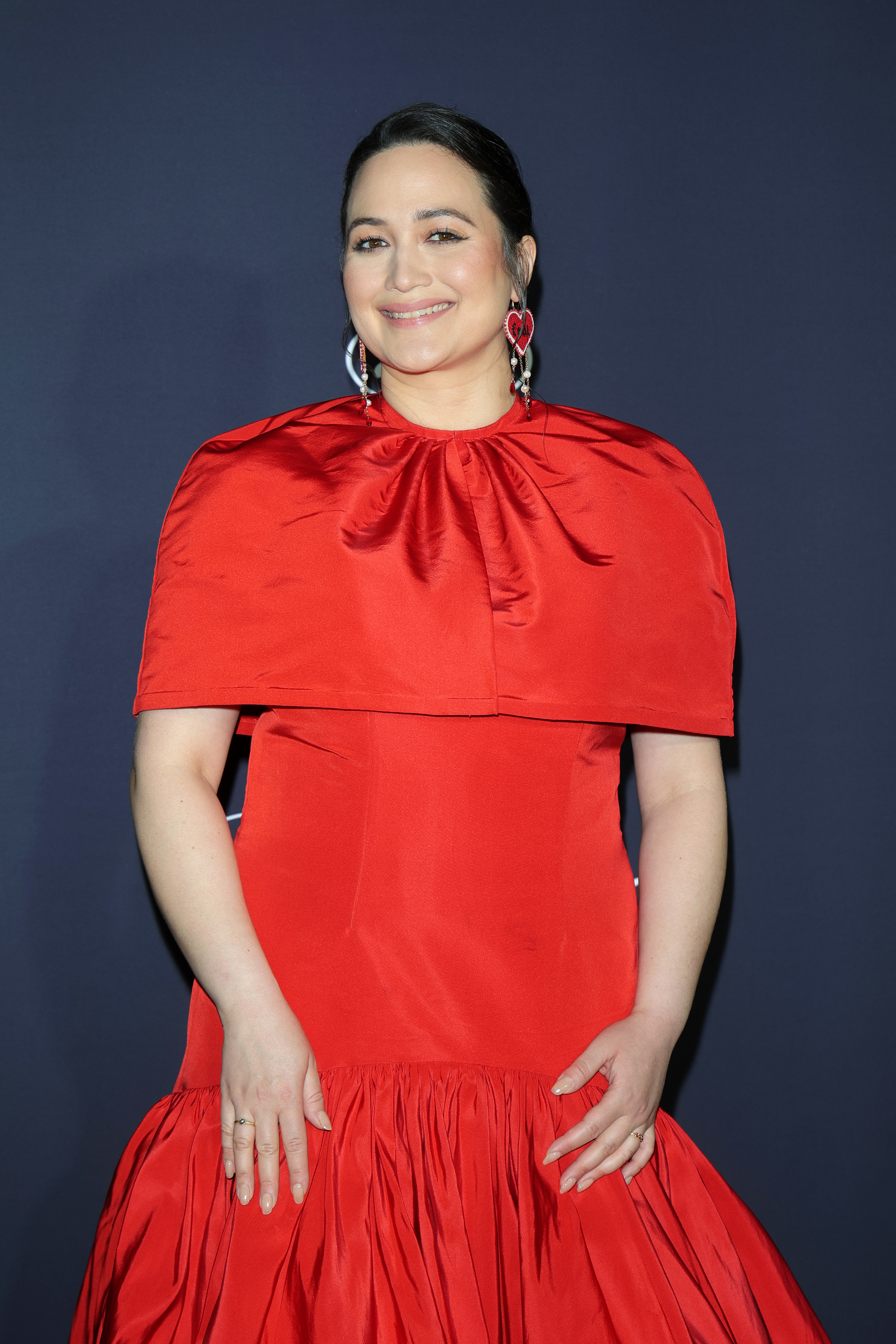 Lily Gladstone wears an elegant red gown with a capelet and matching red earrings at a celebrity event. She smiles at the camera with hands relaxed