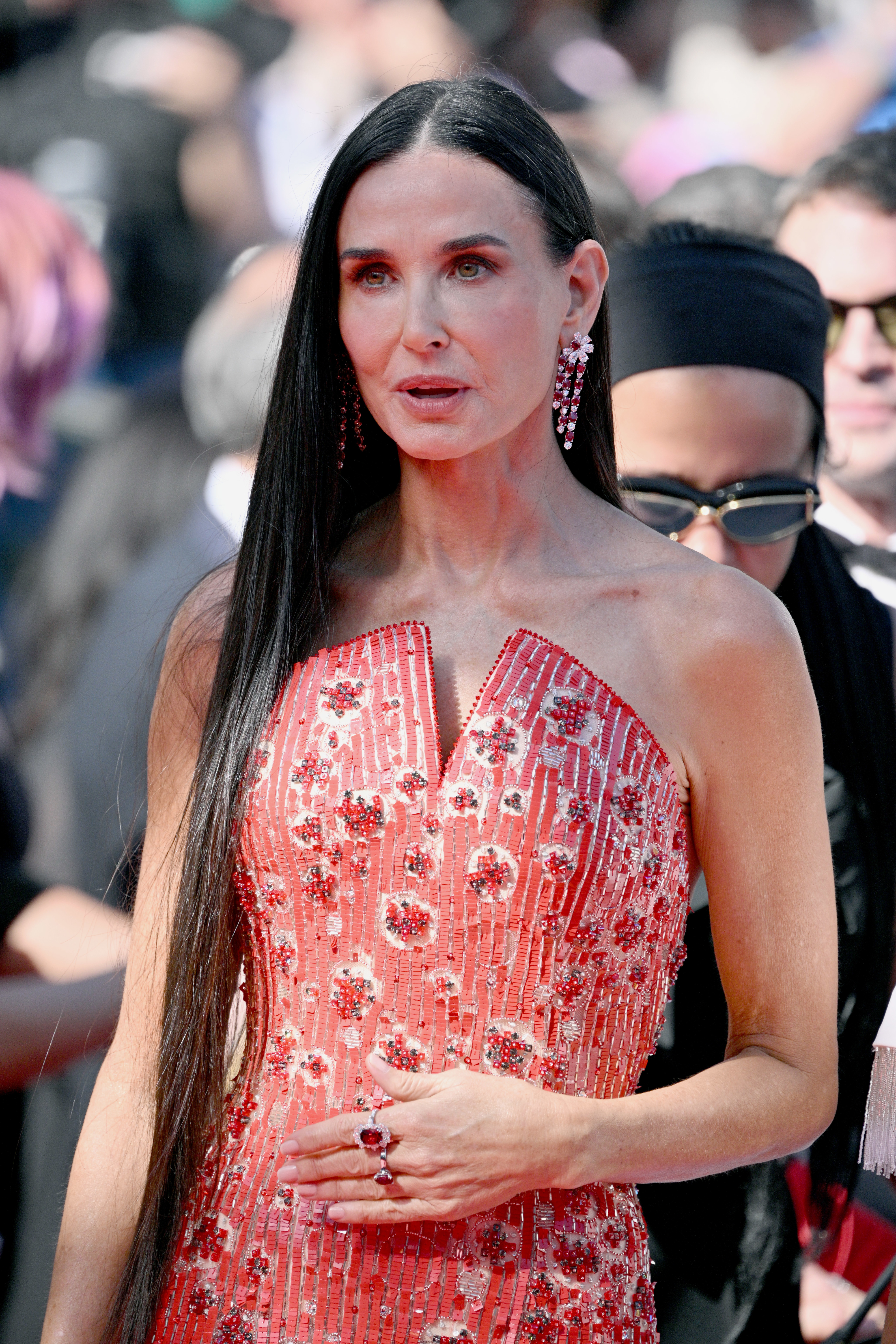 Demi Moore poses on the red carpet in a strapless, sequined gown with floral embellishments. She wears long, straight hair and accessorizes with drop earrings