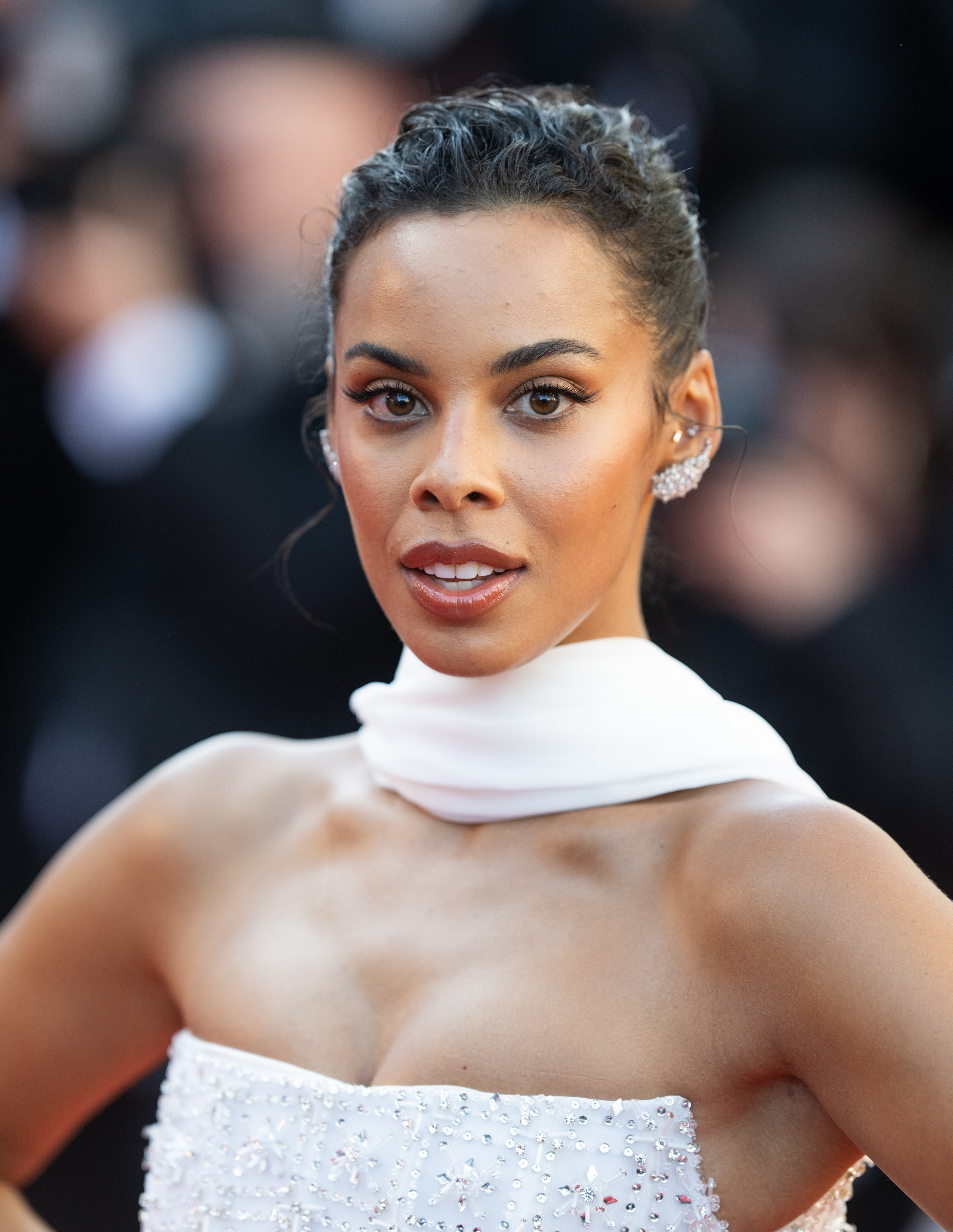 Rochelle Humes poses on the red carpet in a strapless, white beaded gown with a matching scarf around her neck. Glossy updo and elegant earrings complete the look