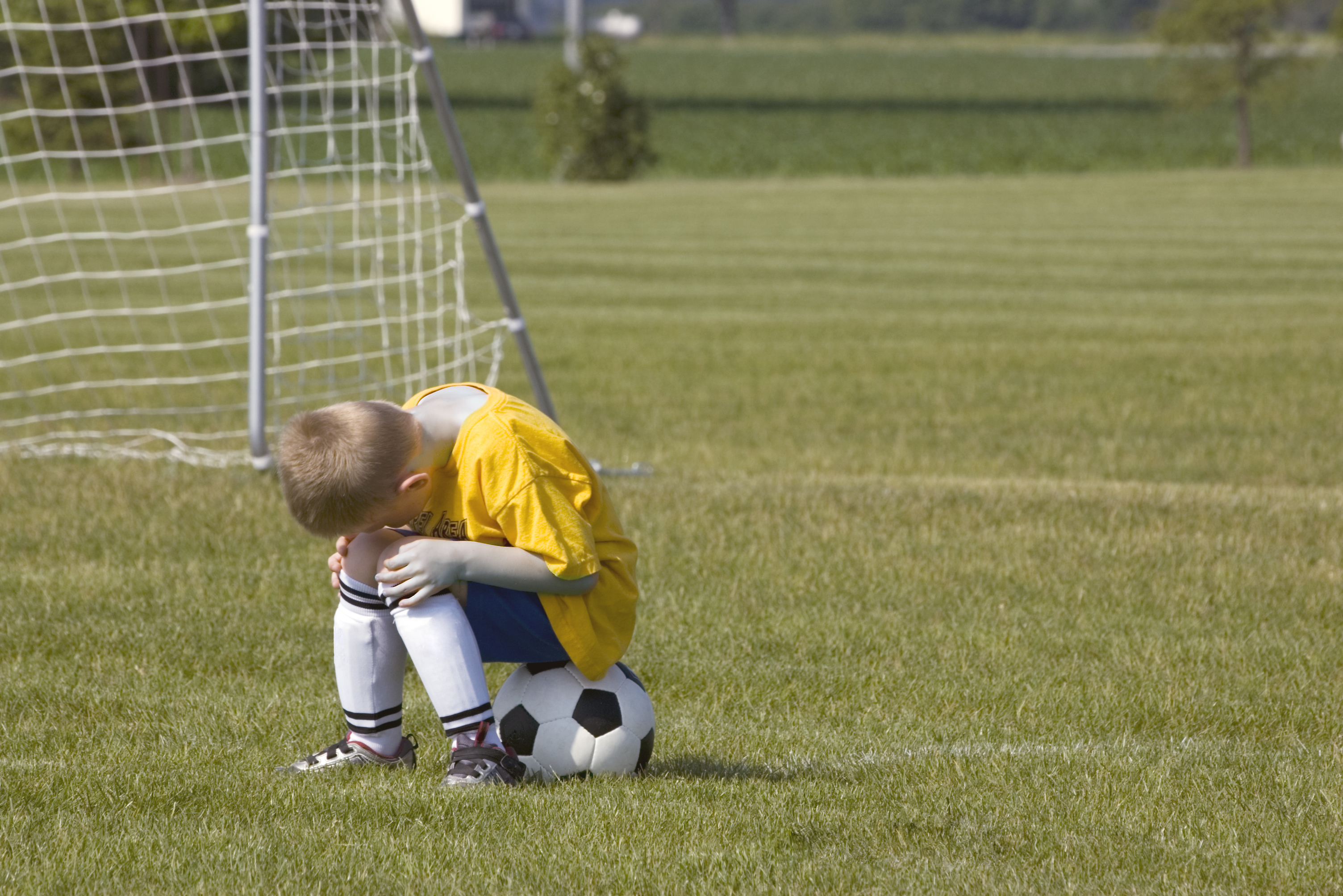 A boy in a soccer uniform sits on a soccer ball on the field, looking down with his head in his hands. The goal net is in the background
