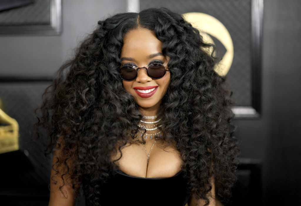 H.E.R. on the red carpet wearing black sunglasses, a black strapless dress, multiple necklaces, and sporting voluminous curly hair at an event