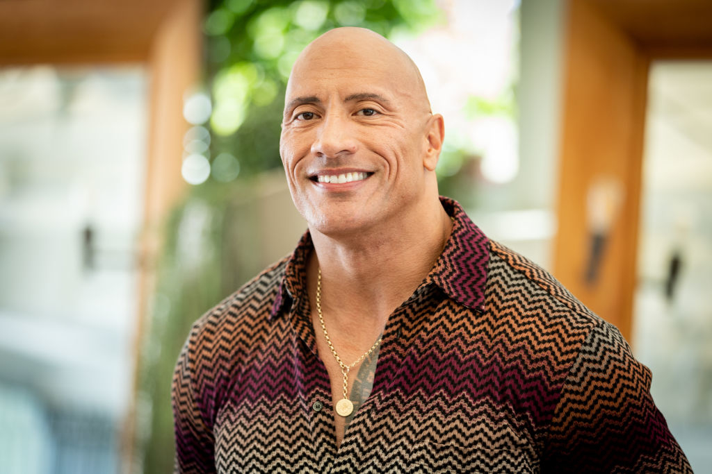 Dwayne Johnson smiling, wearing a patterned button-up shirt with a gold necklace in an indoor setting