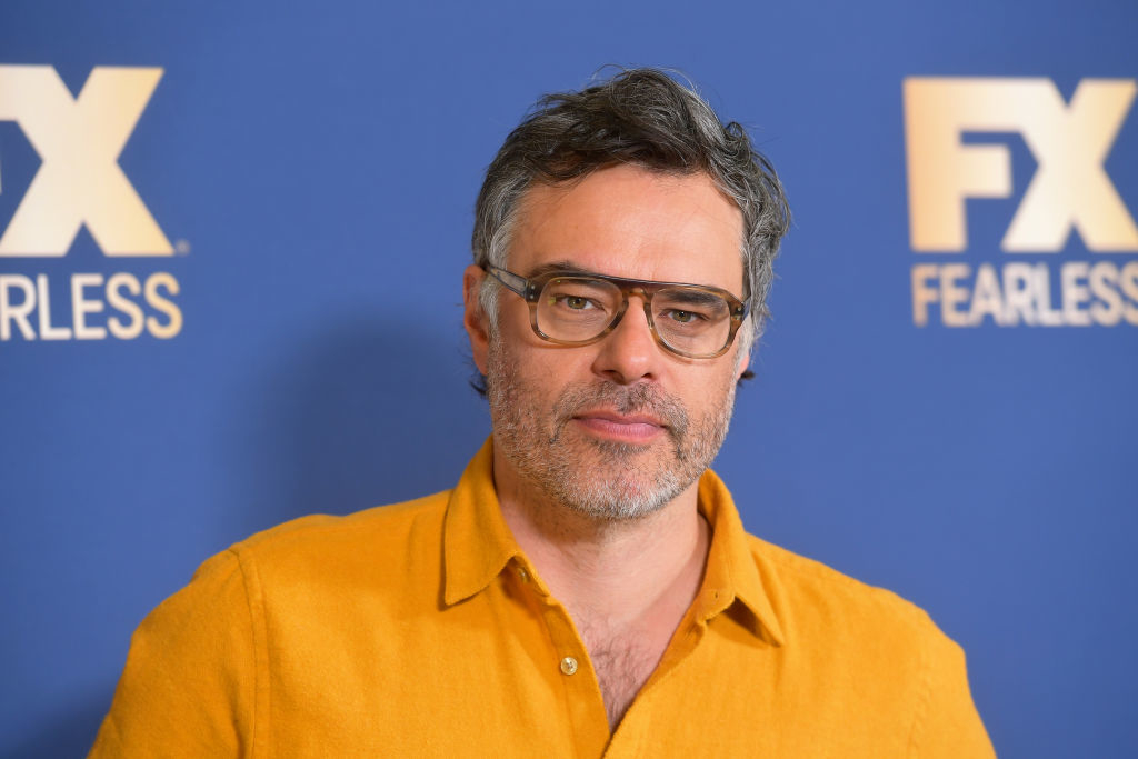 Jemaine Clement stands in front of an FX network backdrop, wearing glasses and a casual button-up shirt