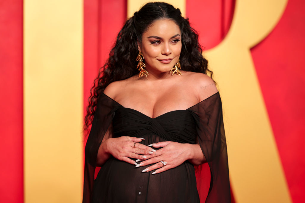 Vanessa Hudgens on the red carpet wearing an off-shoulder black sheer dress, touching her midsection, and sporting gold leaf earrings
