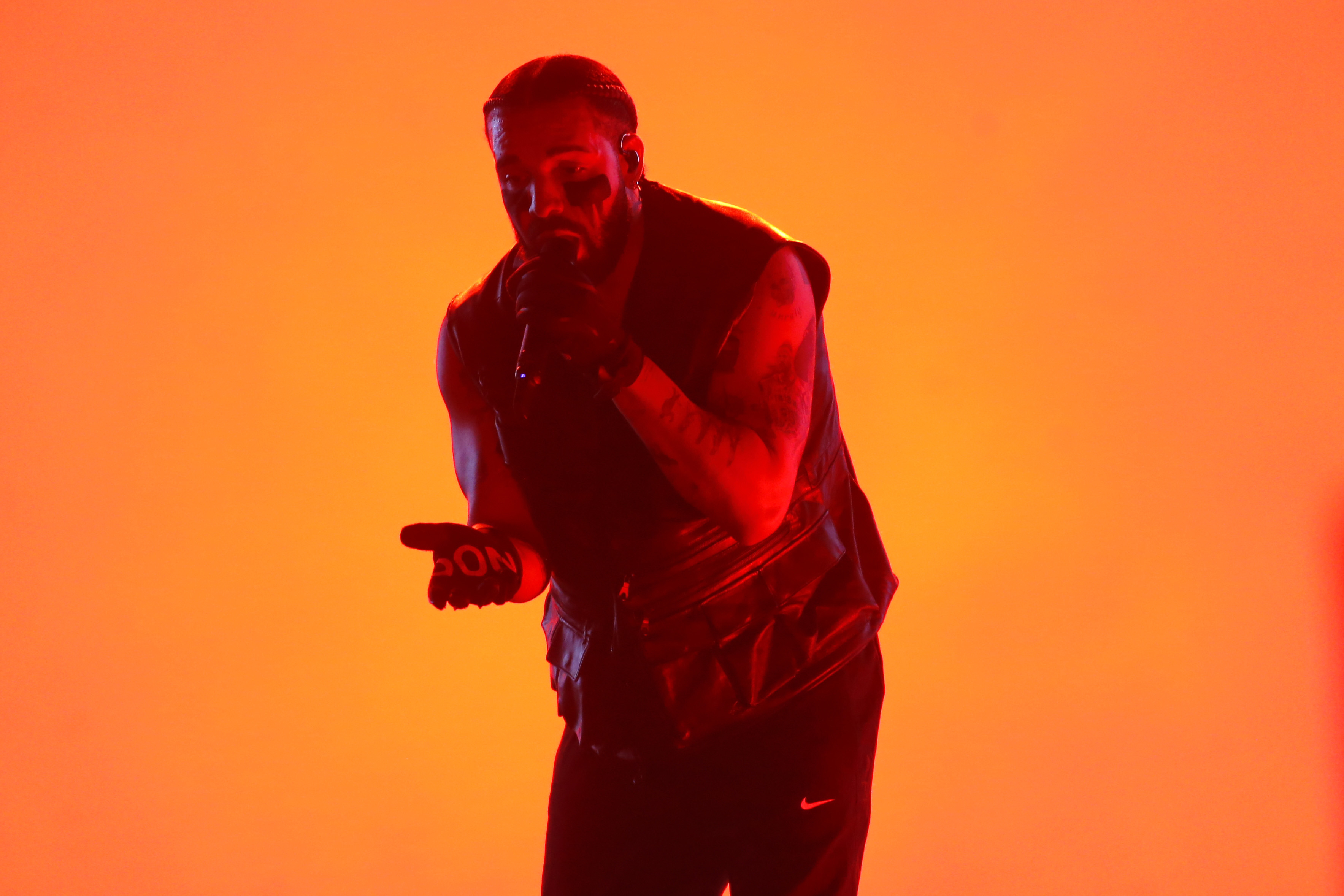 Drake performs on stage with intense red lighting, wearing a sleeveless vest, black gloves, and pants
