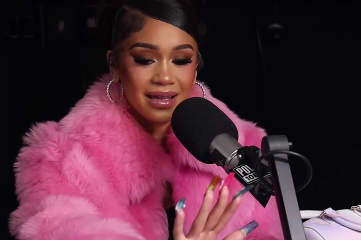 Saweetie, wearing a fluffy coat, talks into a radio microphone during an interview. She gestures with her hand, showcasing her long, colorful nails