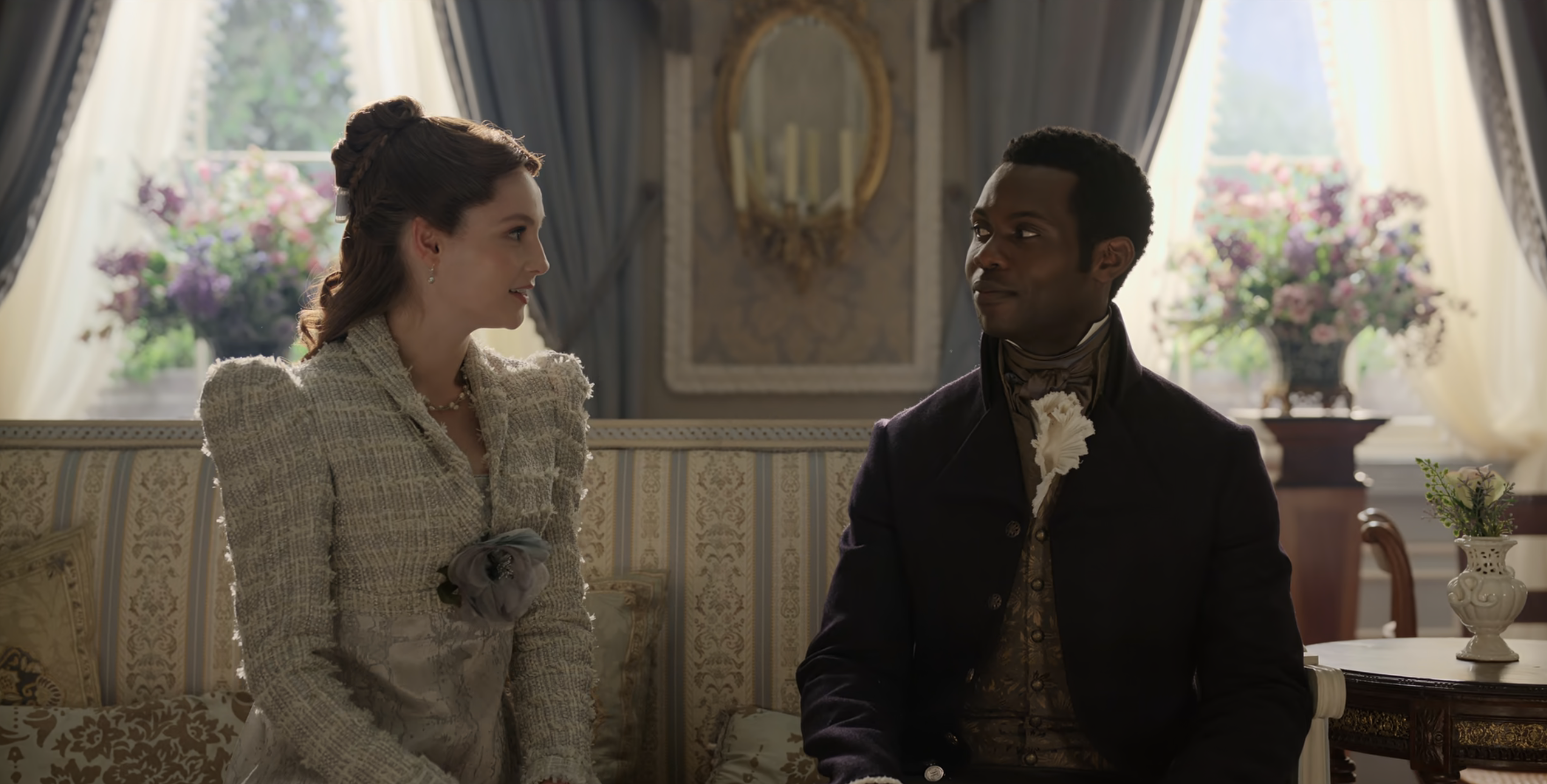 Two characters from Bridgerton are seated on a luxurious couch in an ornate room. The woman wears an elegant dress; the man sports a vintage suit and cravat