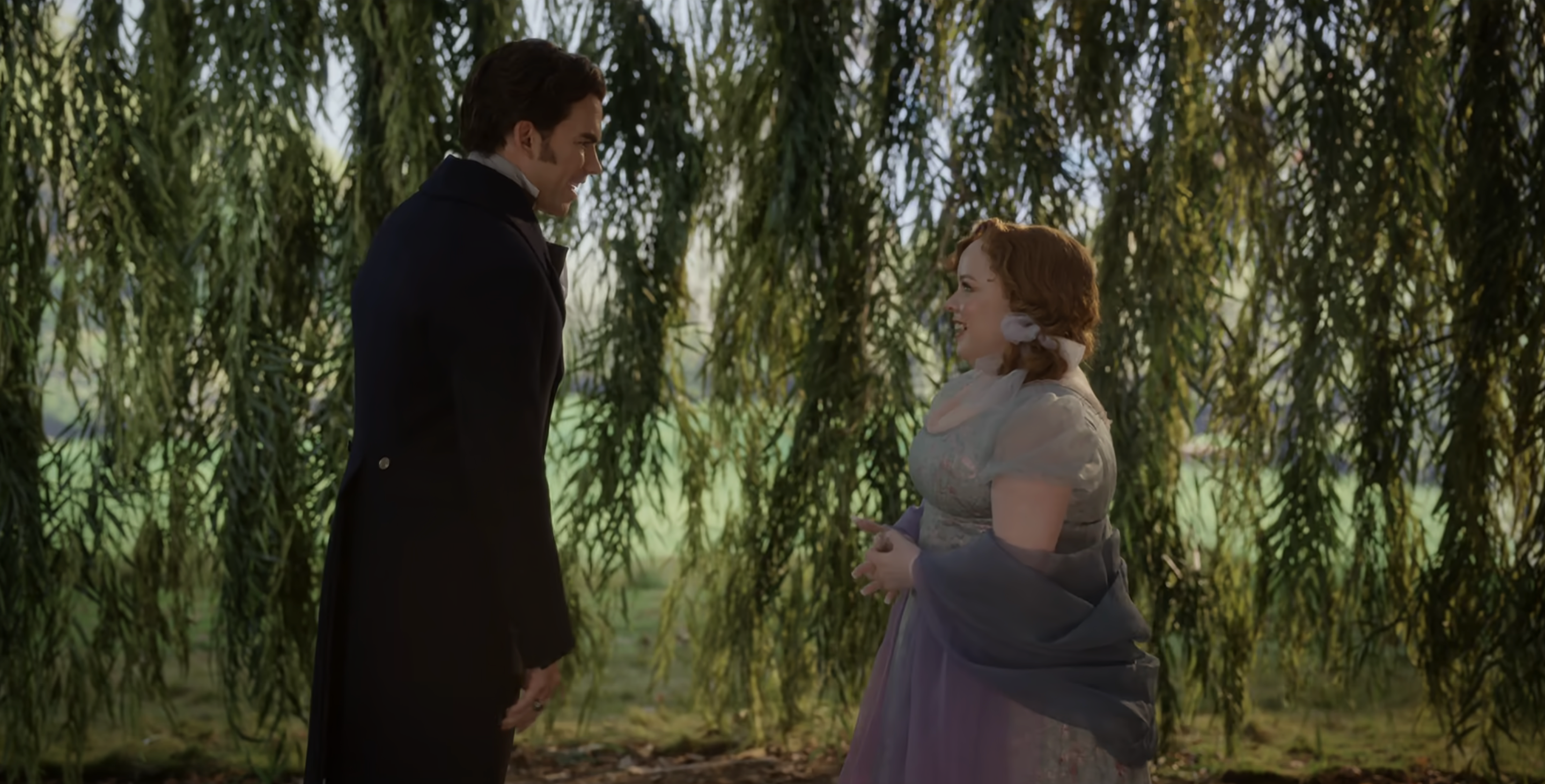 Luke Newton and Nicola Coughlan in period costumes stand facing each other in front of a weeping willow tree