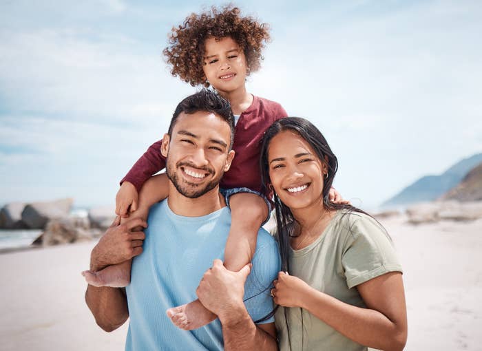 A happy family at the beach with a child on the father's shoulders, both parents smiling at the camera