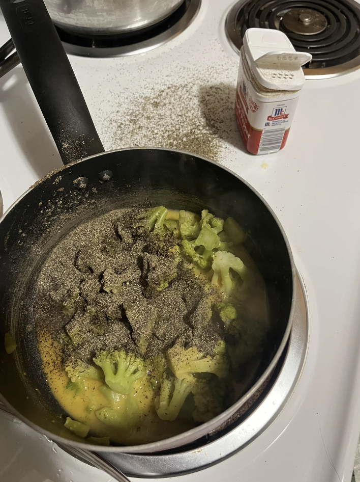 A pan on the stove with broccoli and a large amount of unidentified seasoning, next to a salt container