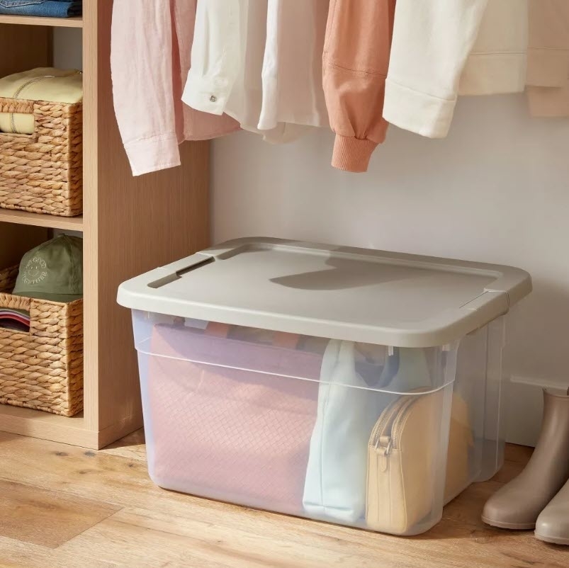 Storage bin with a lid, under hanging clothes and beside boots, used for organizing clothing items
