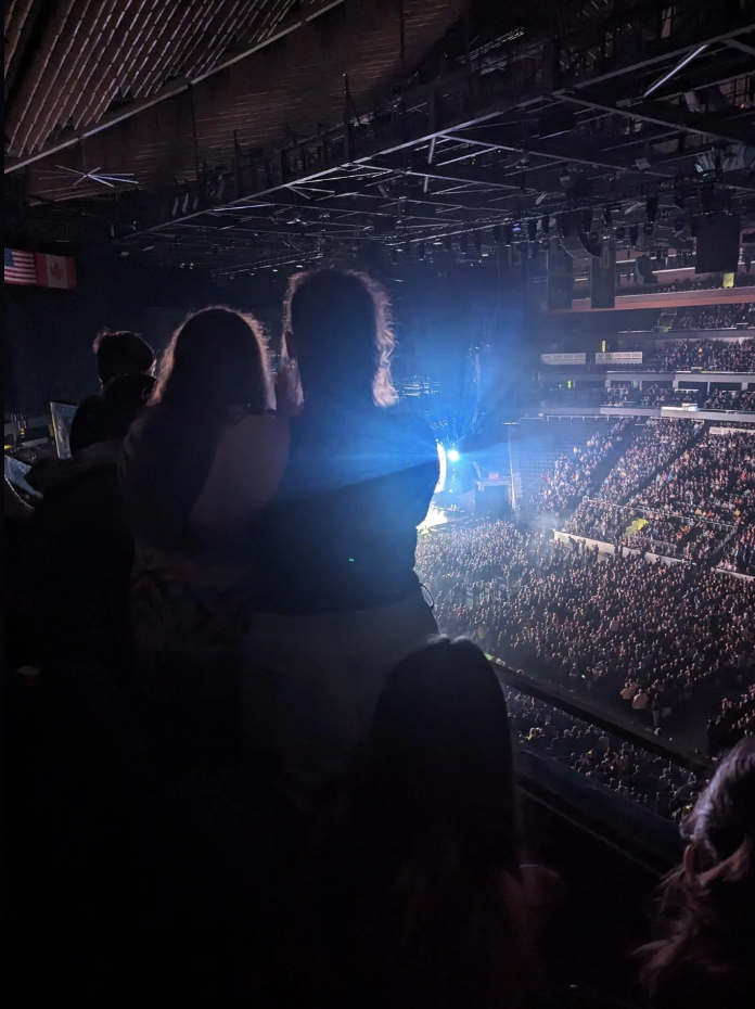 Audience members facing a brightly lit stage at a concert venue, some standing