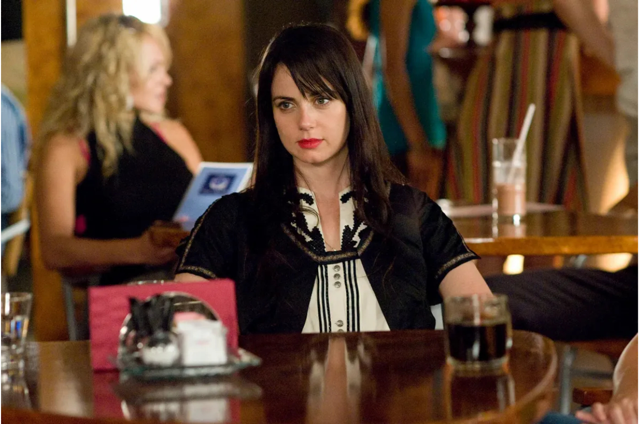 Actress in a black outfit with embellishments sits at a bar in a TV show scene
