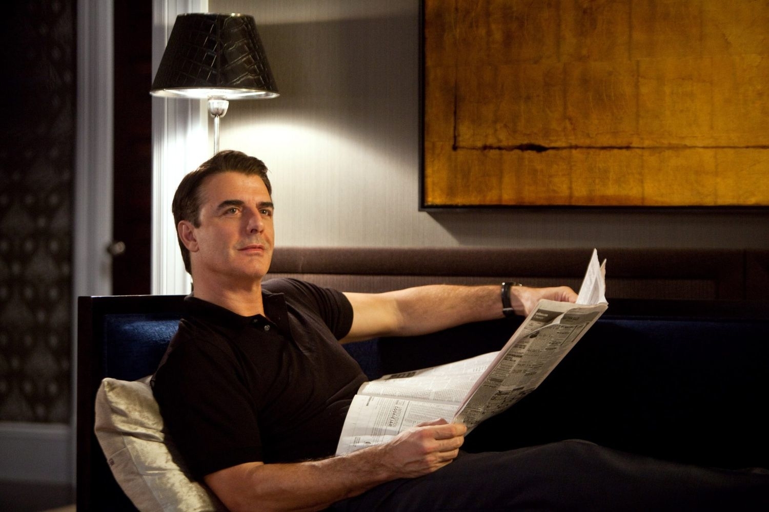 Chris Noth seated, casually dressed, reading a newspaper in a well-lit room