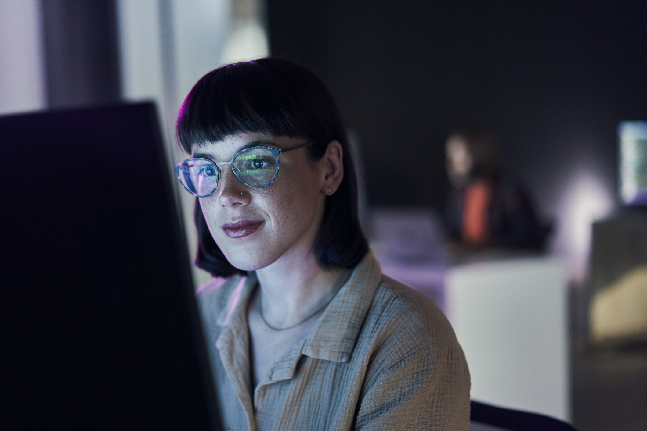 Woman with glasses smiling, working on a computer in an office setting