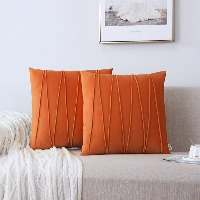 Two pleated orange throw pillows on a sofa with a cozy blanket, near a framed picture