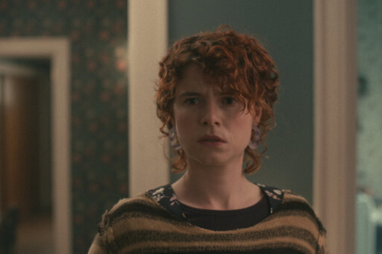 Person with curly hair standing indoors, looking concerned, perhaps a scene from a book adaptation