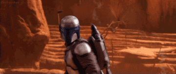 Character in Mandalorian armor walking in a desert-like setting from the TV series &quot;The Mandalorian.&quot;