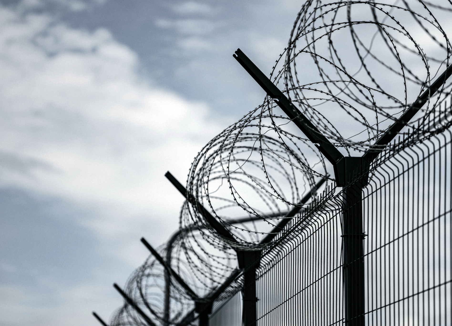 Barbed wire tops a security fence against a cloudy sky, suggesting a secure or restricted area