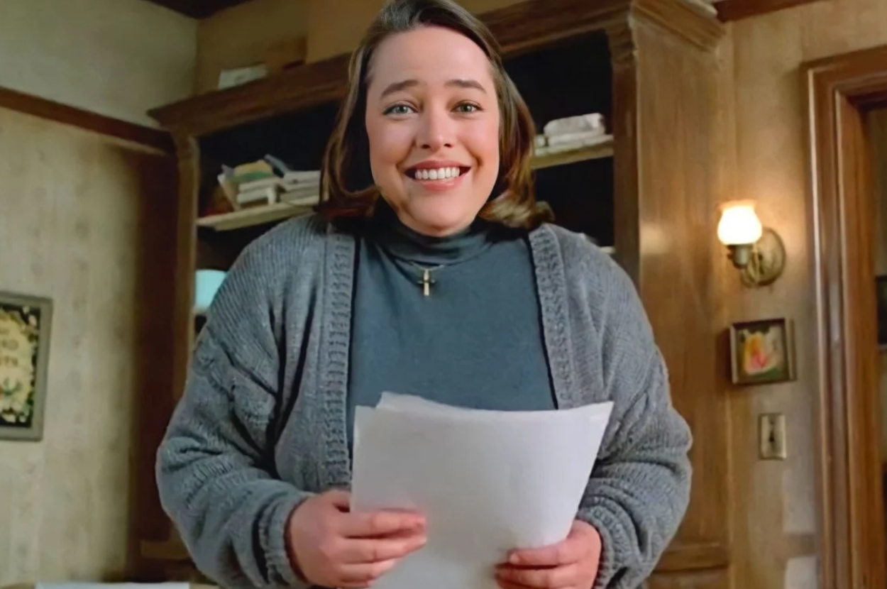 Woman smiling, holding papers, wearing a sweater over collared shirt indoors