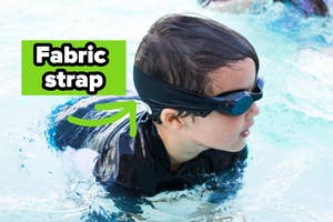 Child in water wearing swim goggles with fabric strap for secure fit. Advertised for comfortable swimming