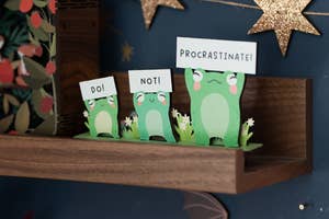 Printed paper frogs on a shelf, one says 'DO!', another 'NOT!', a third 'PROCRASTINATE!'