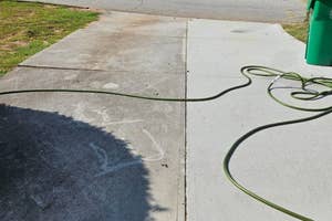 A green garden hose strewn across a concrete sidewalk next to a green trash can