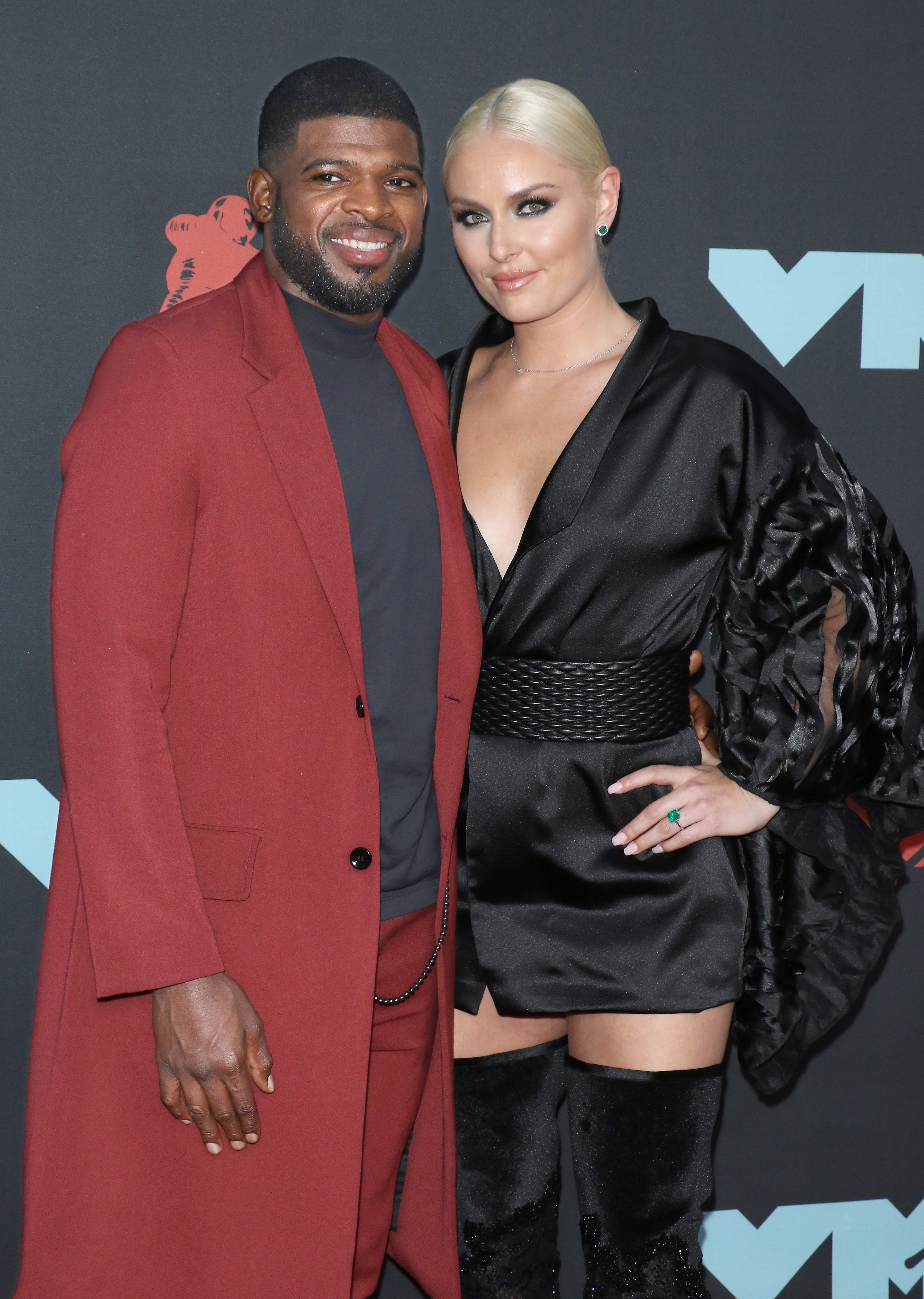 Two celebrities posing together; man in a red coat and woman in a black dress with ruffled sleeve