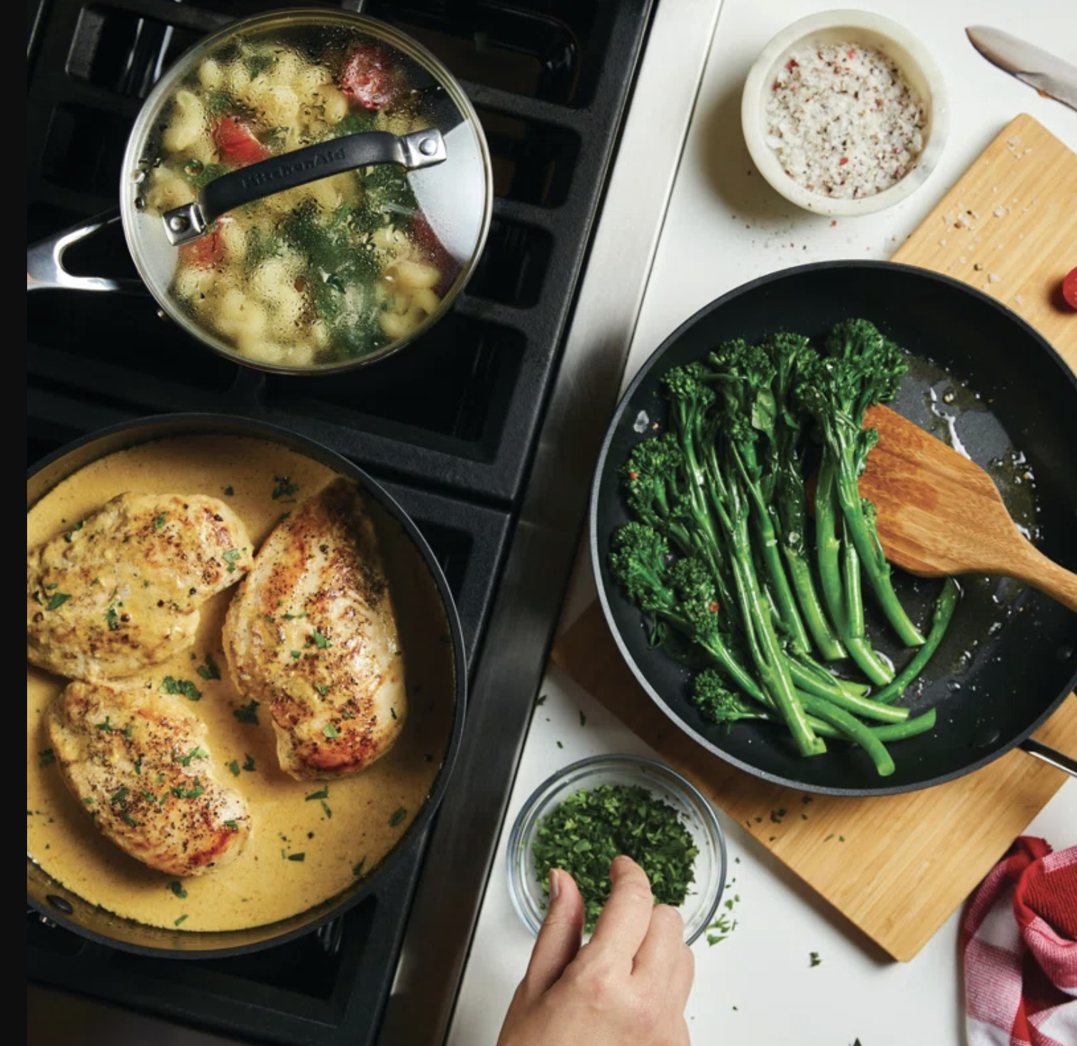 Top-down view of two stovetop pans, one with chicken in sauce, another with sautéed broccoli, next to chopped herbs and seasonings
