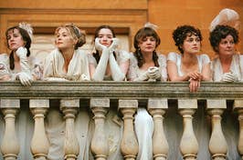 Five women in historical attire with feathered headpieces lean on a balustrade