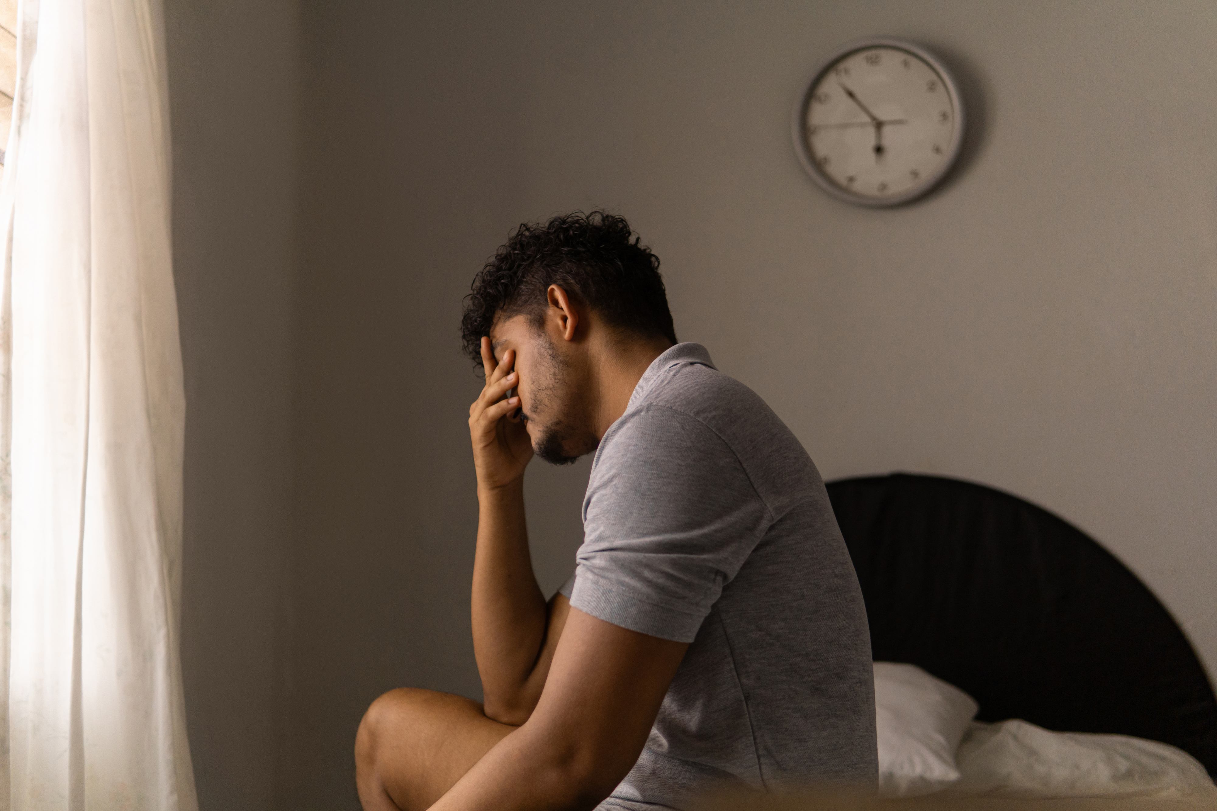 Person sitting on bed with hand on face in a contemplative or distressed pose, clock on the wall in the background
