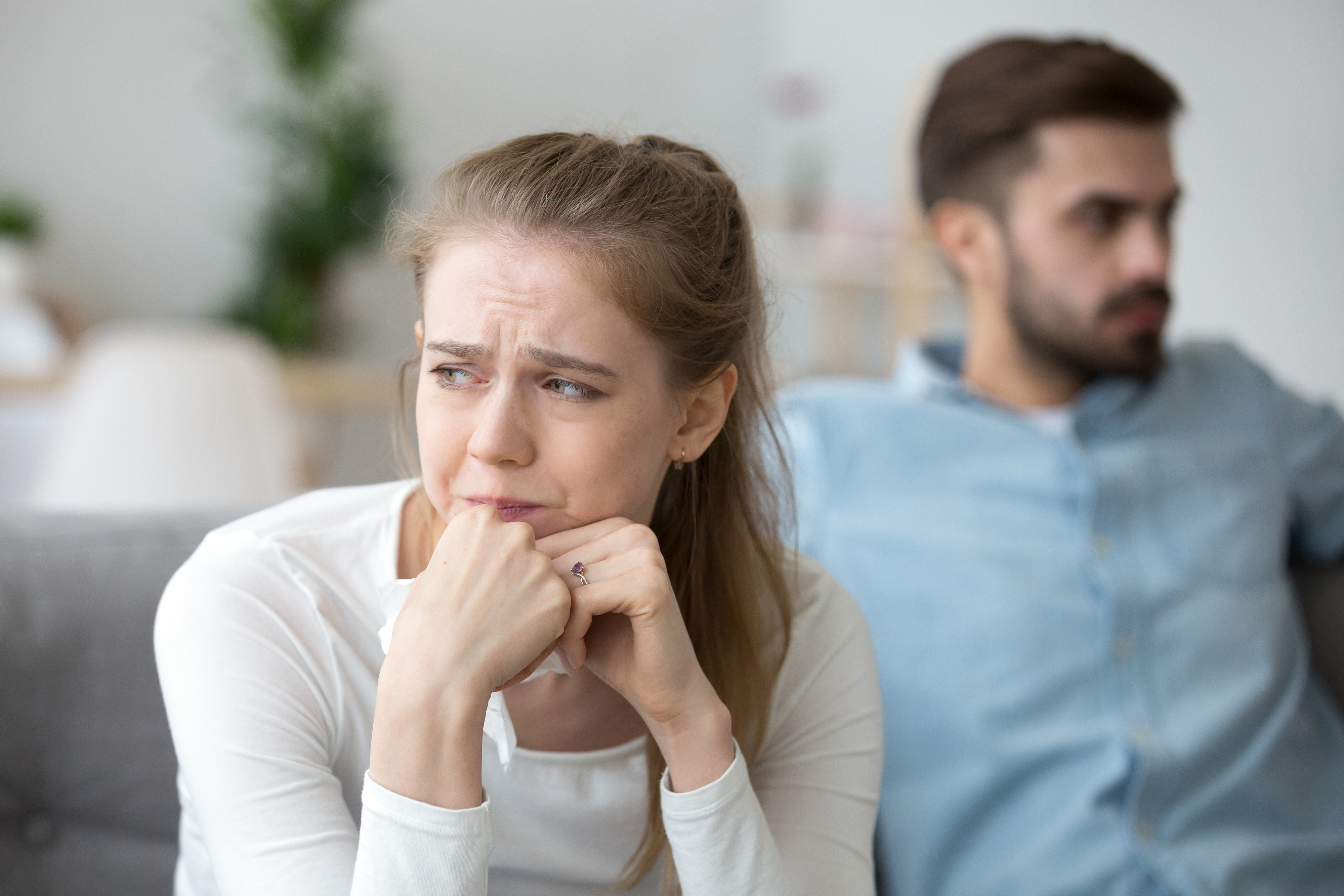 Woman looks anxious, man sits behind her with a blurred background. They appear to be in a disagreement or a stressful situation