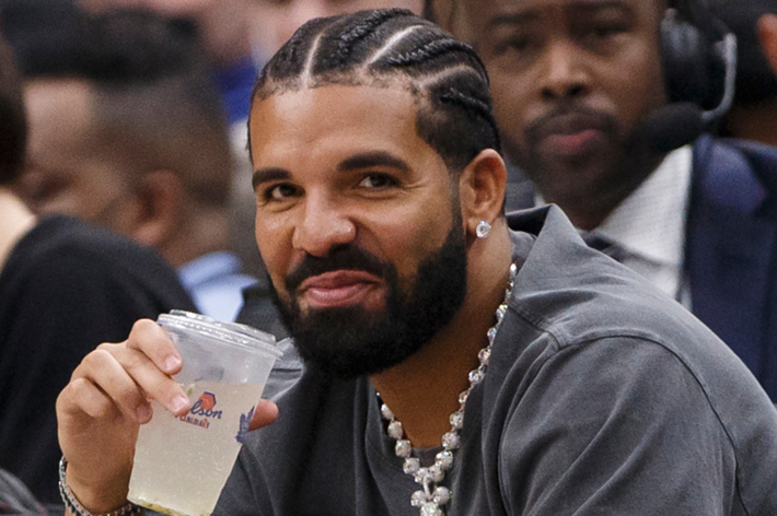 Drake sitting courtside at a basketball game, smiling, wearing a grey outfit and diamond earrings